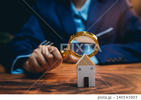Businessman Examining Miniature House with Magnifying Glass in Office Setting for Real Estate Analysis Businessman Examining Miniature House with Magnifying Glass in Office Setting for Real Estate Analysis 140543903