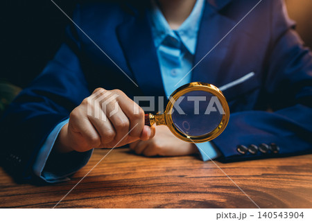 Person Holding Golden Magnifying Glass While Investigating Subjects on Wooden Table in Deep Focus Person Holding Golden Magnifying Glass While Investigating Subjects on Wooden Table in Deep Focus 140543904