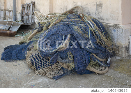 Commercial fishing nets in blue and green colors are piled up against a rustic concrete wall at a busy harbor. The area shows signs of activity with possible nearby boats and workers. Commercial fishing nets in blue and green colors are piled up against a rustic concrete wall at a busy harbor. The area shows signs of activity with possible nearby boats and workers. 140543935