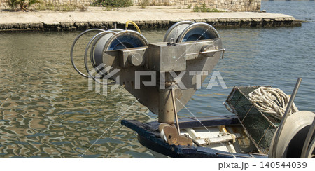 A hydraulic net hauler drum is seen on the bow of a fishing boat. The boat is docked in a port. The water reflects the sunlight and shows a calm harbor environment. 140544039