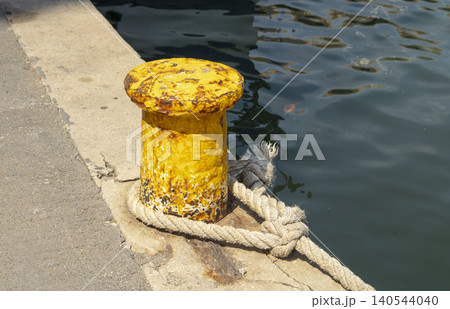 A yellow mooring bollard stands on a concrete pier by the sea. Thick rope is tied around it, ready to secure fishing boats. The water reflects the harbor area. 140544040