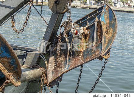A trawl door hangs over water from an industrial fishing boat. Chains and pulleys are visible, showing tools used in maritime work. The scene takes place in a port with boats around. A trawl door hangs over water from an industrial fishing boat. Chains and pulleys are visible, showing tools used in maritime work. The scene takes place in a port with boats around. 140544042