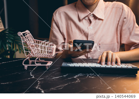 Young person shopping online with smartphone and keyboard, small shopping cart on marble table, modern lifestyle Young person shopping online with smartphone and keyboard, small shopping cart on marble table, modern lifestyle 140544069