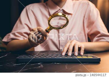 Person Holding Magnifying Glass While Typing on Keyboard in a Modern Workspace Setting Person Holding Magnifying Glass While Typing on Keyboard in a Modern Workspace Setting 140544117