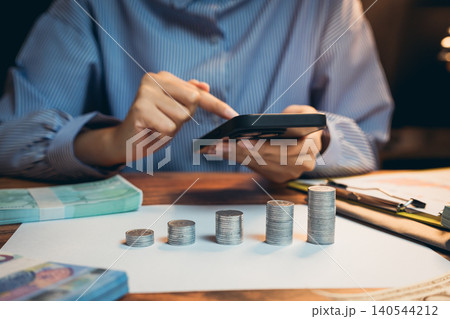 Business concept. Person Analyzing Financial Data Using Calculator with Stacked Coins and Banknotes on Wooden Desk Business concept. Person Analyzing Financial Data Using Calculator with Stacked Coins and Banknotes on Wooden Desk 140544212
