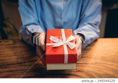 Gift concept. Person Wearing Blue Stripes Shirt Holding Red Gift Box with White Ribbon on Wooden Table Background 140544220