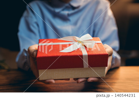 Gift concept. Warm Gesture of Giving with a Red Gift Box in Hands Amidst Cozy Indoor Atmosphere and Soft Lighting Gift concept. Warm Gesture of Giving with a Red Gift Box in Hands Amidst Cozy Indoor Atmosphere and Soft Lighting 140544231