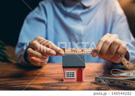 The concept of buying a house. Person Measuring Small House Model with Tape Measure in Home Office Setting, Concept of Real Estate and Design The concept of buying a house. Person Measuring Small House Model with Tape Measure in Home Office Setting, Concept of Real Estate and Design 140544282