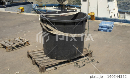 A large black plastic container is placed on a wooden pallet at a commercial shipping dock. It is secured with cargo straps. The harbor is busy with fishing boats nearby. A large black plastic container is placed on a wooden pallet at a commercial shipping dock. It is secured with cargo straps. The harbor is busy with fishing boats nearby. 140544352