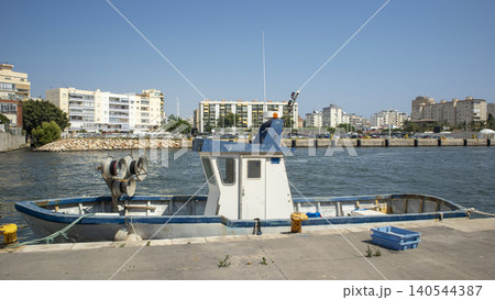 A small blue and white commercial fishing boat is tied at a concrete pier in a busy harbor. The surrounding area features several buildings and a calm water view. A small blue and white commercial fishing boat is tied at a concrete pier in a busy harbor. The surrounding area features several buildings and a calm water view. 140544387