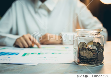 Concept of saving money. Detailed Analysis of Business Growth with Coins in Glass Jar on Wooden Table at Night Concept of saving money. Detailed Analysis of Business Growth with Coins in Glass Jar on Wooden Table at Night 140544399