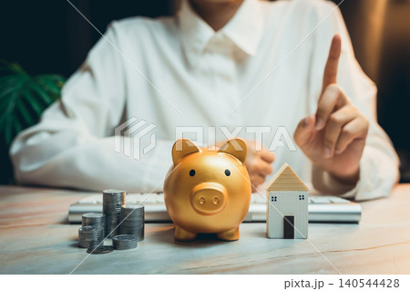 Home buying concept. Woman in White Shirt Points Finger While Golden Piggy Bank and House Model Are Placed on Table with Coins 140544428