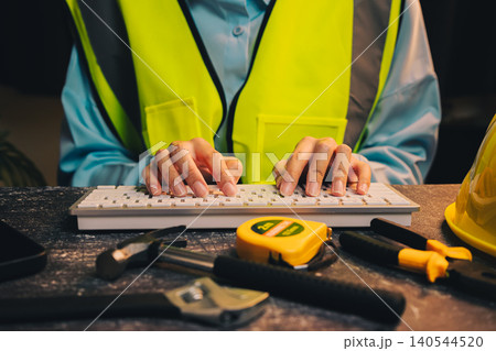 Engineer concept. Construction Worker in Safety Vest Typing on Keyboard Surrounded by Tools and Equipment for Project Management Engineer concept. Construction Worker in Safety Vest Typing on Keyboard Surrounded by Tools and Equipment for Project Management 140544520
