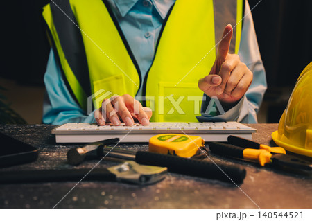 Engineer concept. Construction Worker in Safety Vest Typing on Keyboard Surrounded by Tools and Equipment Engineer concept. Construction Worker in Safety Vest Typing on Keyboard Surrounded by Tools and Equipment 140544521