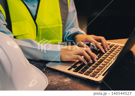 Engineer concept. Construction Worker in Safety Vest Using Laptop at Workplace with Hard Hat on Desk Engineer concept. Construction Worker in Safety Vest Using Laptop at Workplace with Hard Hat on Desk 140544527