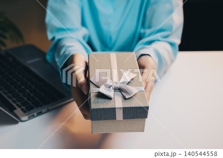 Gift concept. Person with gift box and ribbon near laptop on white table in cozy indoor environment 140544558