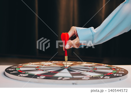 Business Target concept. Hand Reaching to Place Red Dart on Colorful Dartboard in Relaxed Indoor Setting Business Target concept. Hand Reaching to Place Red Dart on Colorful Dartboard in Relaxed Indoor Setting 140544572