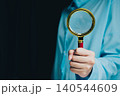 Businessman Holding Magnifying Glass in Blue Shirt with Dark Background for Inspection and Investigation Purposes 140544609