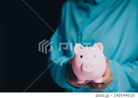 Businessman Holding Pink Piggy Bank Against Dark Background for Saving Money and Financial Planning Concept Businessman Holding Pink Piggy Bank Against Dark Background for Saving Money and Financial Planning Concept 140544610