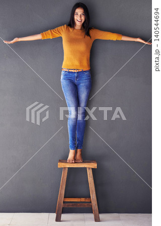 Woman, confident and stand on chair in portrait, smile and fashion on wall background. Female person, clothes and happy lady on stool, apparel and trends in living room at home in house 140544694