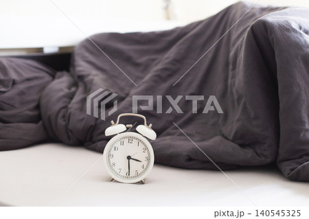 White alarm clock sitting on bed with grey blanket White alarm clock sitting on bed with grey blanket 140545325