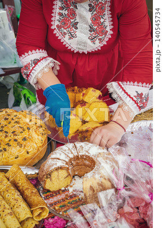 Woman in embroidered dress cuts traditional bread and cake at Kazanluk Rose Festival in Bulgaria. Festival food display features kozunak, banitsa, baking, heritage, community, Bulgarian cuisine Woman in embroidered dress cuts traditional bread and cake at Kazanluk Rose Festival in Bulgaria. Festival food display features kozunak, banitsa, baking, heritage, community, Bulgarian cuisine 140545734