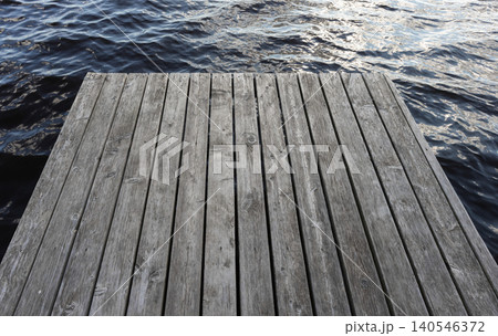 A weathered gray wooden dock extends into calm water, highlighting textured planks A weathered gray wooden dock extends into calm water, highlighting textured planks 140546372