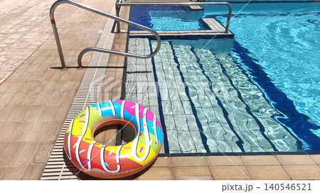 inflatable donut ring float with pink frosting print and multicolor sprinkle pattern on pool deck beside hotel pool, full donut resting on tiled poolside in summer 140546521