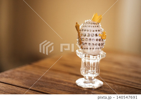 Decorative Easter egg adorned with rhinestones and a small crown, displayed on a crystal pedestal against a softly blurred background Decorative Easter egg adorned with rhinestones and a small crown, displayed on a crystal pedestal against a softly blurred background 140547691