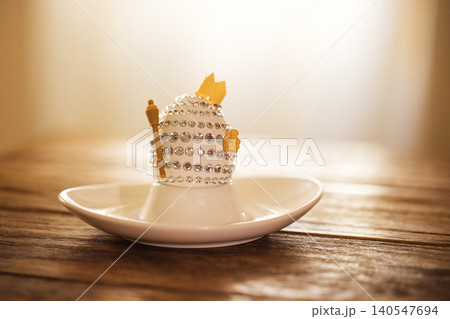Decorative egg-shaped ornament adorned with rhinestones and a golden crown, placed on a white saucer atop a rustic wooden table with soft lighting in the background Decorative egg-shaped ornament adorned with rhinestones and a golden crown, placed on a white saucer atop a rustic wooden table with soft lighting in the background 140547694