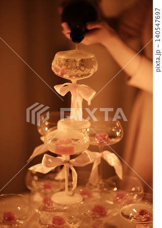 Champagne tower being filled with sparkling wine, decorated with pink flowers and white ribbons, illuminated by soft warm light in a festive setting Champagne tower being filled with sparkling wine, decorated with pink flowers and white ribbons, illuminated by soft warm light in a festive setting 140547697