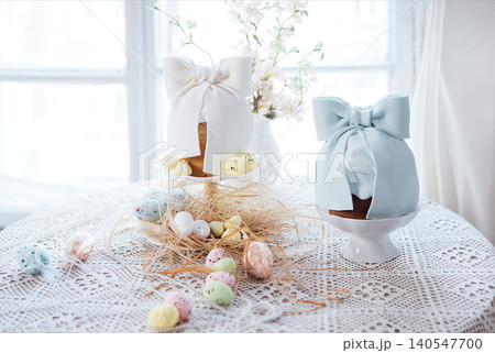 Decorative Easter cakes with pastel bows on a lace tablecloth, surrounded by colorful eggs and floral accents in a bright indoor setting 140547700