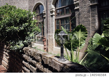 Stone wall with a vintage lamp post beside lush green plants and large leaves, featuring tall windows of a brick building in the background 140548384