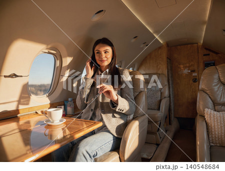 Caucasian young adult woman talking on phone and holding glasses inside a private jet cabin 140548684