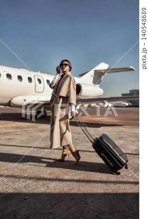Stylish Caucasian mid adult woman talking on phone pulling suitcase on airport tarmac Stylish Caucasian mid adult woman talking on phone pulling suitcase on airport tarmac 140548689