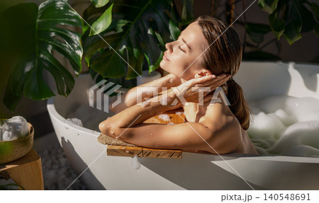 Caucasian young woman relaxing in a bubble bath at home spa with green plants Caucasian young woman relaxing in a bubble bath at home spa with green plants 140548691