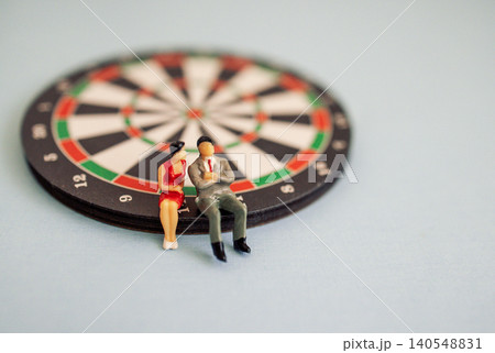 Miniature figurines of a man and woman sitting on a dartboard, with a soft blue background creating a playful and whimsical scene Miniature figurines of a man and woman sitting on a dartboard, with a soft blue background creating a playful and whimsical scene 140548831