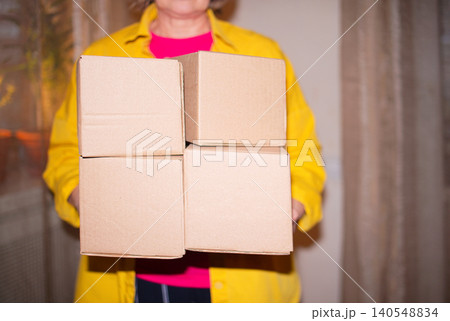 Woman in yellow shirt holds four stacked cardboard boxes in front of a window with sheer curtains, indoor setting with soft lighting and a warm atmosphere Woman in yellow shirt holds four stacked cardboard boxes in front of a window with sheer curtains, indoor setting with soft lighting and a warm atmosphere 140548834