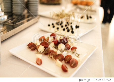Assorted appetizers featuring skewered grapes and cheese cubes on white plates, elegantly arranged for a catering event with additional dishes in the background 140548892