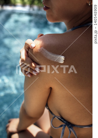 Woman applying sunscreen on her shoulder while sitting by a swimming pool, with water and greenery visible in the background Woman applying sunscreen on her shoulder while sitting by a swimming pool, with water and greenery visible in the background 140548909