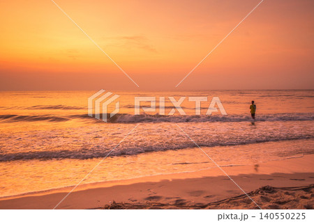 Lone figure wading into the ocean at sunrise with warm golden light reflecting on the water 140550225