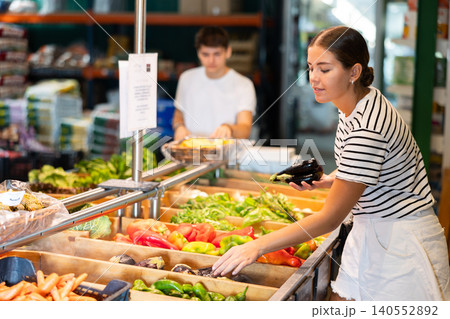 Positive girl choosing fresh eggplants in vegetables department of supermarket 140552892