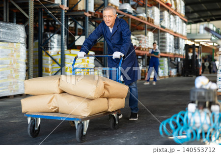 In warehouse rack area of store, loader mature man pushes and carries large cart for bulky cargo In warehouse rack area of store, loader mature man pushes and carries large cart for bulky cargo 140553712