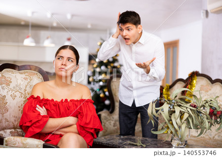 Displeased young European man and woman in red dress yelling at each other on sofa during preparations for New Year in living room Displeased young European man and woman in red dress yelling at each other on sofa during preparations for New Year in living room 140553746