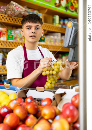 Young male supermarket worker weighing on the scales green grapes in grocery shop Young male supermarket worker weighing on the scales green grapes in grocery shop 140553764