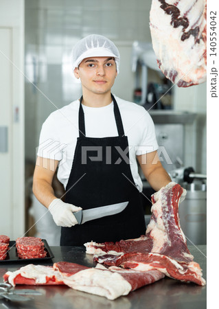 Guy employee of butcher shop cuts meat carcass into small pieces of meat for retail sale in store 140554042