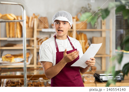 Cheerful young baker studying financial documents about profit in artisan bakery 140554140