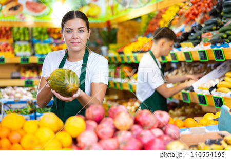 Positive young saleswoman holding fresh melon during offering fresh fruits Positive young saleswoman holding fresh melon during offering fresh fruits 140554195