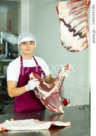 Guy employee of butcher shop cuts meat carcass into small pieces of meat for retail sale in store Guy employee of butcher shop cuts meat carcass into small pieces of meat for retail sale in store 140554198
