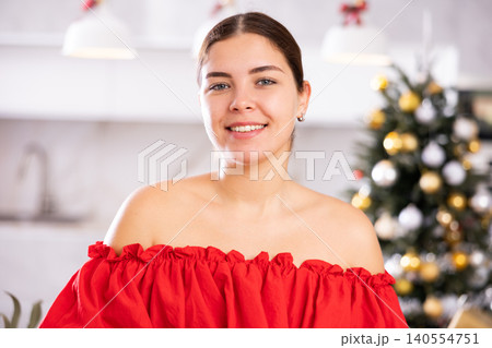 Portrait of happy young female in red dress smiling at camera in front of decorated Christmas tree at home Portrait of happy young female in red dress smiling at camera in front of decorated Christmas tree at home 140554751
